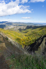 Bare rocks descend into a gorge. Geology and rock layers are visible. Mountains, hills and a sky with clouds in the background