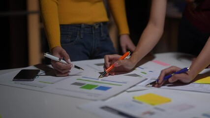 Two people are working on a project together, with one person holding a cell phone and the other holding a pen. Scene is collaborative and focused