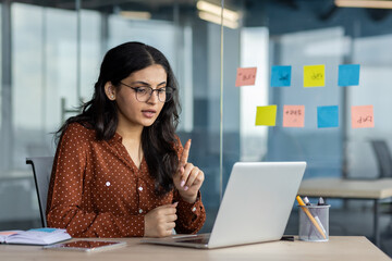 Wall Mural - Hispanic woman businesswoman participating in virtual meeting on laptop. Displays professional communication, focus, and remote work in modern office. Ideal for business and technology concepts.