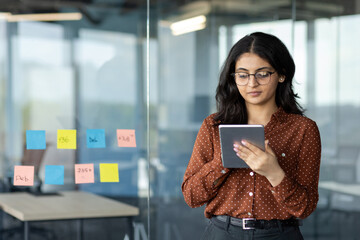 Wall Mural - Hispanic woman businesswoman using digital tablet in office. Focused and organized, planning projects with colorful sticky notes on glass wall, showcasing effective business strategy.