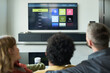 © pressmaster - Rear view of diverse couple and child watching television screen in modern living room, with remote in hand and streaming options visible on screen