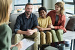 © pressmaster - Multiracial family sitting on couch during counseling session, smiling and holding hands. Professional counselor in foreground with clipboard in hand and tablet on table