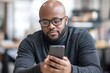 © ChaoticMind - A bald man with a beard and glasses is deeply engaged with his smartphone, showing a profound connection with technology in a modern, blurred office setting.