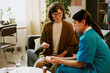© AnnaStills - Healthcare professional assisting elderly woman with medication instruction, senior woman seated on sofa in a cozy room with plants and furniture