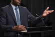 © Seventyfour - Cropped shot of African American male politician dressed in blue suit gesticulating with hands speaking to audience, while giving public speech on stage at podium against black curtains