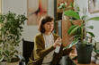 © AnnaStills - Woman watering houseplants in a living room filled with greenery, contemporary art on walls, and various decorative pieces creating a cozy home environment