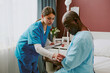 © AnnaStills - Nurse providing medical care to senior black patient sitting in hospital room with healing equipment and technology around treatment focused on patients arm