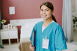 © AnnaStills - Smiling nurse wearing medical scrubs standing in patient's room while holding clipboard in hands looking ready for healthcare duties