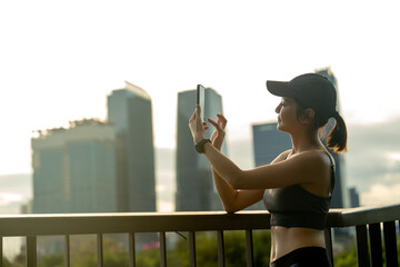 Wall Mural - Asian sport woman use mobile phone to take photo and relax after exercise in public garden with high building of big city in background and soft warm light.