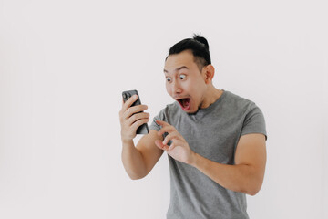 Wall Mural - Shock and wow face Asian man in grey t-shirt use smartphone app on white.