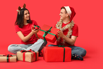 Wall Mural - Happy young couple with Christmas tinsel and gift boxes on red background