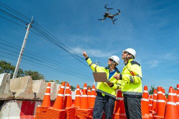 two engineers in high visibility jackets and helmets operate a drone using a laptop amidst a stack o