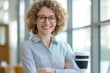 © Vooglo.com - Professional woman curly hair glasses confident smile arms crossed beige blazer white shirt office background