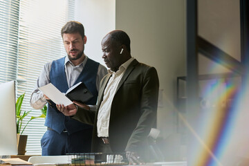 Poster - Young male economist with open notepad explaining data on paper document held by mature African American colleague