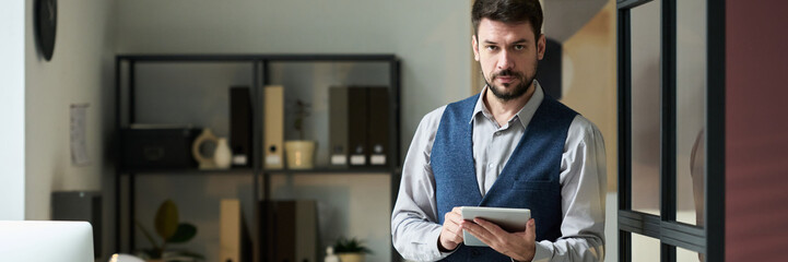 Poster - Header of young serious businessman using tablet and looking at camera while standing in coworking space with desks and shelves