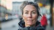 © iuricazac - The image features a middle-aged woman with a warm smile sporting messy brown hair with gray streaks and a black jacket with a red lining stands in front of a blurred city street