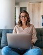 © Marko - Happy young woman working on laptop sitting on sofa in living room at home. Businesswoman in casuals working from home