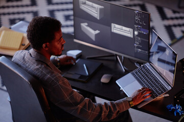 Fototapete - African American man working with virtual 3D models on dual computer screens in dark room with focused lighting. Using modern gadgets and design software with attention to detail