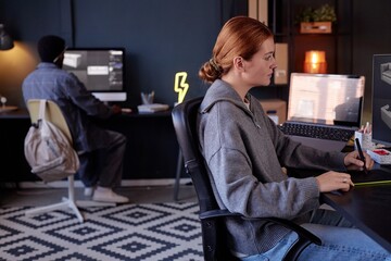 Fototapete - Woman sitting at desk with digital tools working on project while colleague at separate desk in modern office environment creating dynamic workspace atmosphere