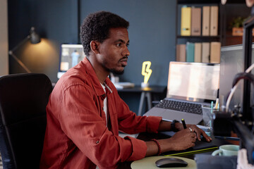 Poster - Man working late in modern office, concentrating on task. Sitting at desk with computer, bookshelves in background, surrounded by office supplies