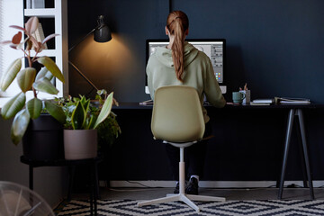 Poster - Red-haired woman working remotely in stylish home office setup, focusing on computer screen displaying graphic design project, lamp providing ambient lighting