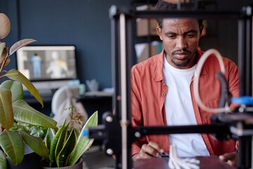 Leinwandbilder - African American man working with 3D printer in modern office space focusing on precise details with computer in background and plants decorating workspace