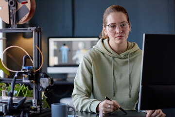 Leinwandbilder - Portrait of woman wearing glasses and gray hoodie sitting at desk with computer and 3D printer in modern office setting