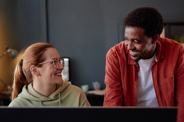 Poster - Smiling colleagues engaged in discussion in a modern office setting. Person with glasses wearing hoodie, while another in red shirt, standing with monitor in background