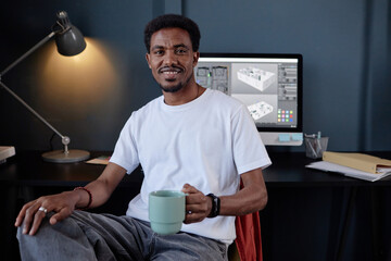 Leinwandbilder - Portrait of cheerful man looking at camera while holding coffee cup, with computer displaying design software in background. Lamp and desk accessories creating an office ambiance