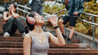 © peopleimages.com - Woman, headphones and drinking water for exercise in park, mineral liquid and fitness recovery. Female person, athlete and nutrition for hydration on stairs, workout break and streaming song in Japan