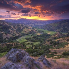 Wall Mural - Vibrant sunset over a valley landscape with rolling hills, green meadows, and rocky foreground.