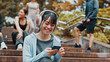 © peopleimages.com - Woman, fitness break and scroll with phone on park stairs with headphones for online exercise program. Sports, cellphone and Japanese athlete with website for workout, routine and outdoor in nature