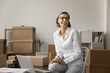 © fizkes - Happy grey haired business owner woman posing in e-commerce storage room, sitting on stepladder at workplace, enjoying Internet store management, looking away, smiling, laughing