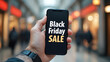 © artmozai - A person holding a phone with the words 'Black Friday Sale' on it in front of a shopping mall background an online marketing concept