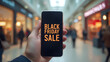 © artmozai - A person holding a phone with the words 'Black Friday Sale' on it in front of a shopping mall background an online marketing concept