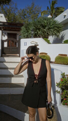 Poster - Young hispanic woman outdoors in lanzarote canary islands wearing sunglasses and a black dress in front of a white house with tropical plants