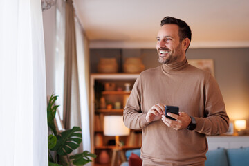 Wall Mural - Positive adult man smiling and looking through window while texting over smart phone in his apartment