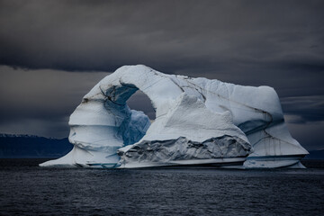 Wall Mural - dark and stormy skies over a blue iceberg that has been carved into an arch by the arctic winds. kai