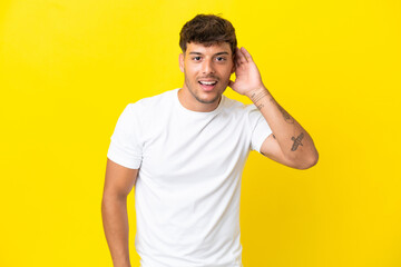 Sticker - Young caucasian handsome man isolated on yellow background listening to something by putting hand on the ear