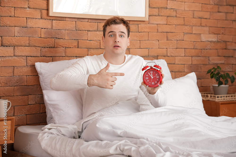 Shocked young man pointing at alarm clock in bedroom