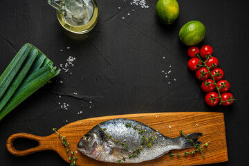 Wall Mural - Fresh, raw dorado fish on a wooden board on a black background with leeks, lime, olive oil and cherry tomatoes on the table ready for cooking. Space for text