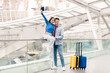 © Prostock-studio - A young couple stands joyfully at the airport, with a suitcase nearby. The woman holds a passport high in the air, while her partner smiles, conveying excitement for their upcoming travel adventure.