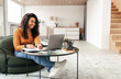 © Prostock-studio - Online Training Concept. Smiling casual black lady sitting at tea coffee table working on pc laptop writing letter in paper notebook, taking notes watching weninar, free copy space, full body length