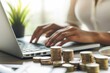 © zakiroff - Businesswoman working on laptop surrounded by stacks of coins, representing financial success and growth in today's business world