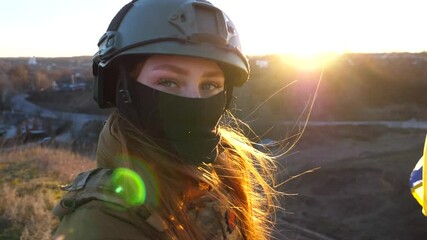 Wall Mural - Portrait of female military medic of ukrainian army looks into camera against waving blue-yellow banner. Girl in camouflage uniform and helmet holding flag of Ukraine at field on sunny day. Slow mo