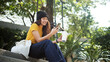 © ChayTee - Gen Z eco go green woman asia sit happy relax at tree public park city life enjoy typing mobile phone drink coffee bottle use reuse tote bag care save the earth planet net zero waste clean future hope