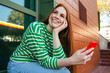 © VISTA by Westend61 - Happy redhead woman sitting with mobile phone near building