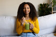 © Daniel - African American woman relaxing at home sitting on the sofa drinking healthy green juice looking at camera.