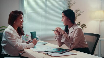 Poster - Two businesswomen engage in a conversation at a desk in an office. One gestures with a pen, emphasizing points while discussing strategies and ideas for their work