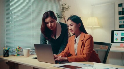 Fototapete - Two businesswomen collaborate in an office at night, analyzing data on a laptop. Engaged in discussion, they strategize and plan for their project, showcasing teamwork and professionalism